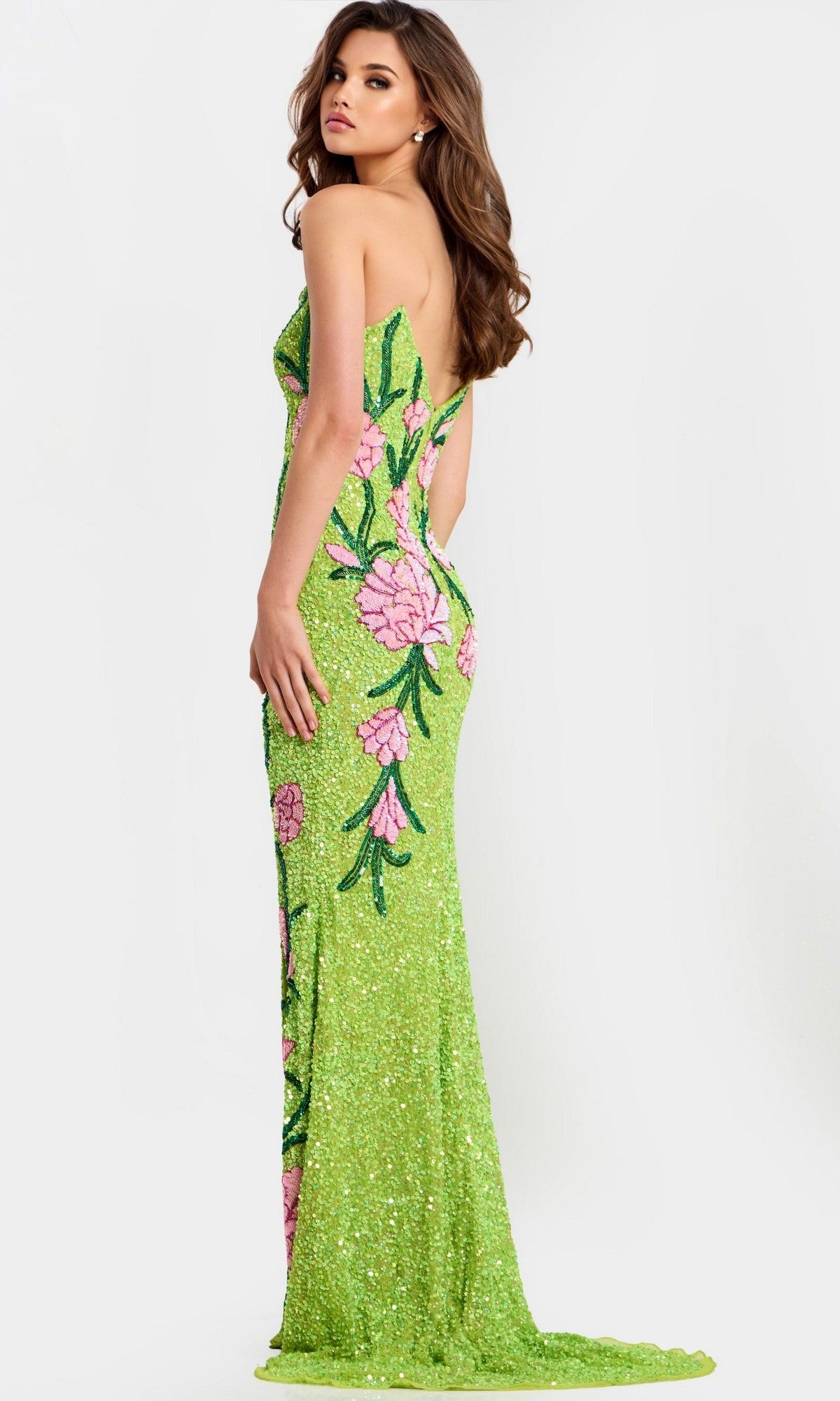 A woman poses with her back and side to the camera against a plain light background, wearing the Jovani 49231 long prom dress in lime green with pink floral designs.